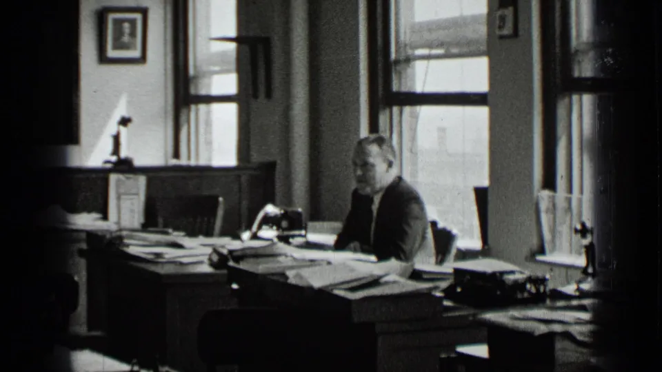 In a 1939 New York office interior, a man in a suit sits at a desk covered with papers, with windows and framed wall art behind him.