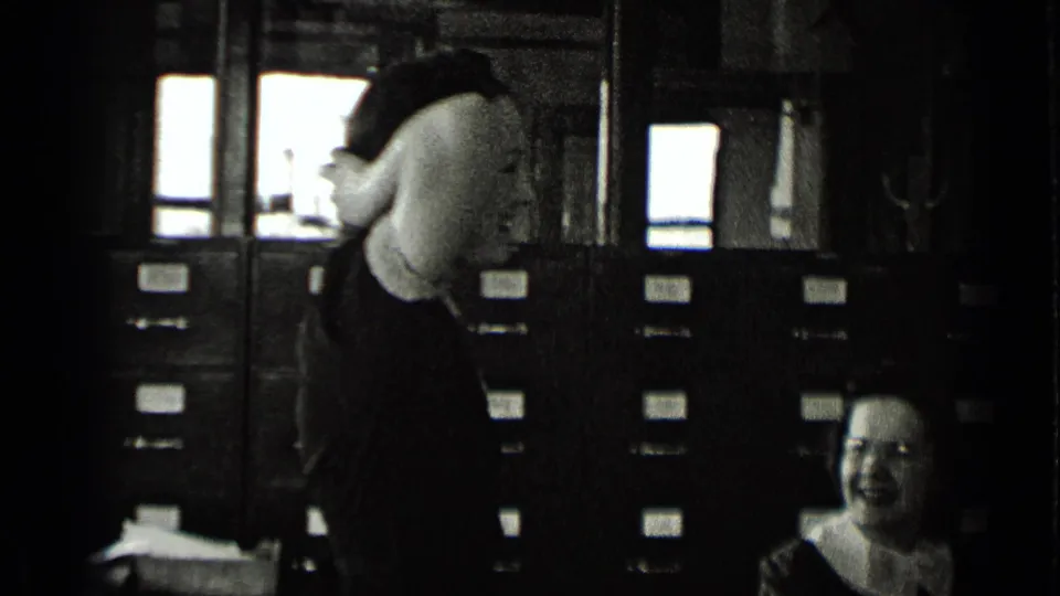 In a New York office filing room, one woman stands near filing cabinets while another seated woman smiles at the lower right.
