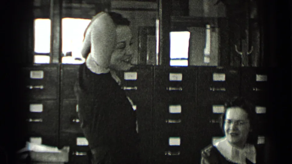 A standing woman adjusts her hair in front of filing cabinets while a seated woman watches in the office filing room.