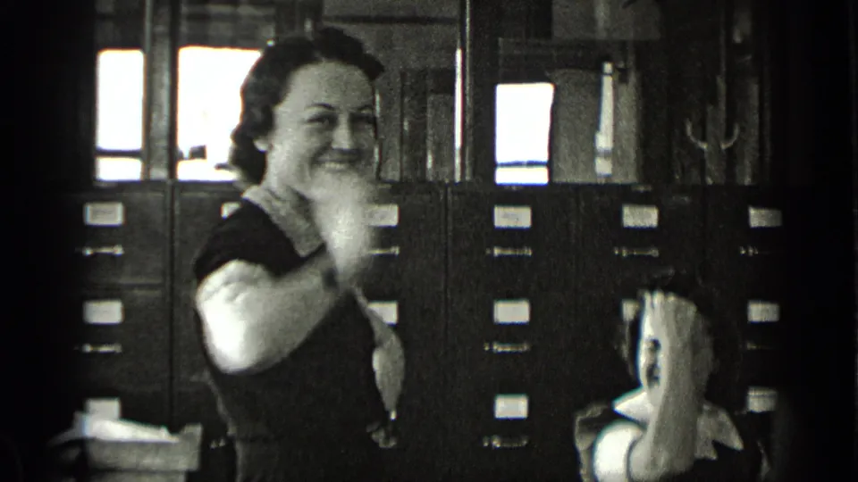 A smiling woman gestures toward the camera as another woman laughs seated beside filing cabinets in the 1939 New York office scene.