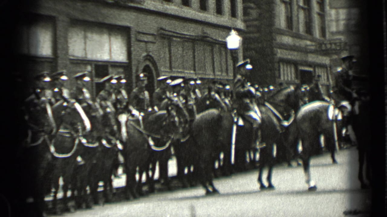 Mounted uniformed riders and horses hold position on a New York street, with a streetlamp and building facade behind them.