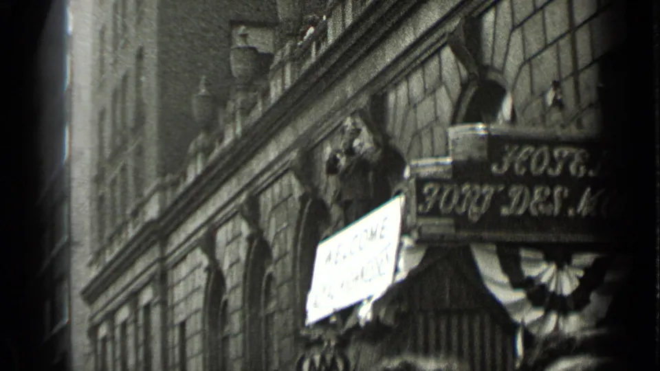 New York street view with a photographer near a building facade and a visible hotel sign, captured in black-and-white 8mm footage.