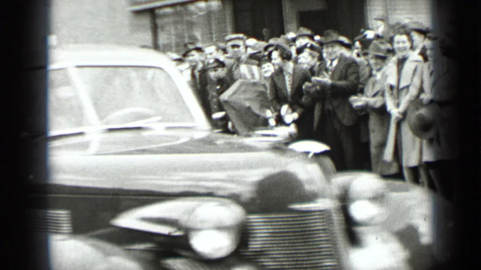 1939 New York street scene with a rounded classic automobile in the foreground and spectators gathered along the sidewalk.
