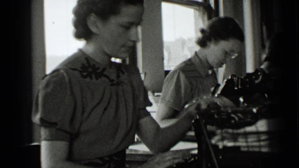 In a New York office setting from 1939, two women sit at desks with manual typewriters and papers near tall windows.