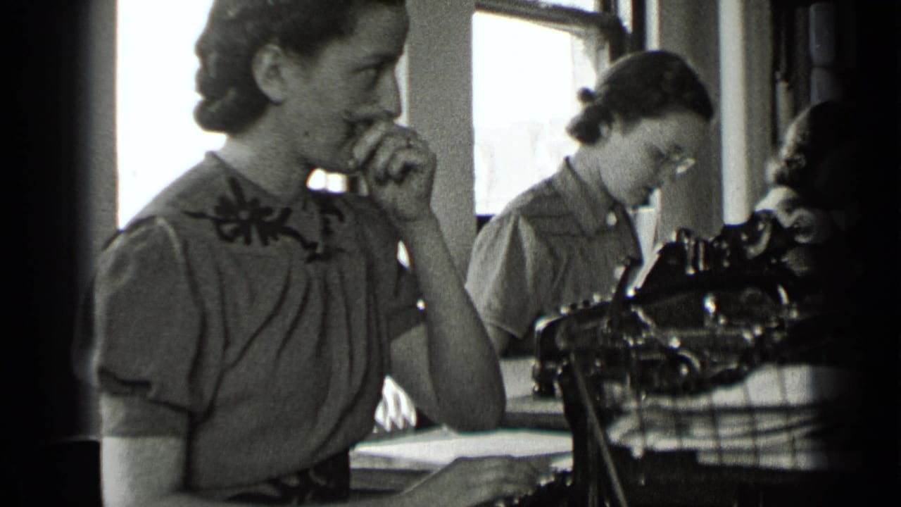 Woman seated by a typewriter touches her nose while another woman works behind her.