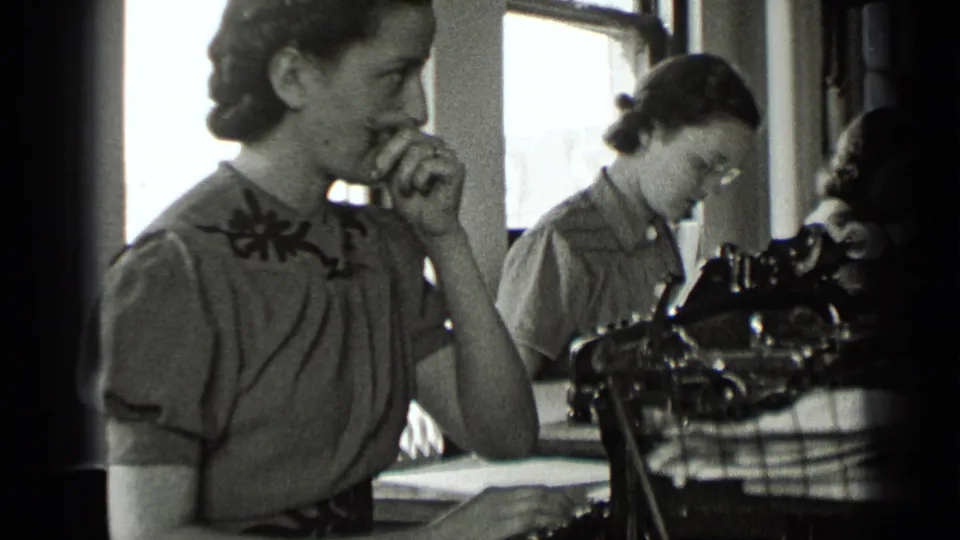 A woman in a 1939 New York office touches or scratches her nose beside a typewriter while another worker looks down at papers.