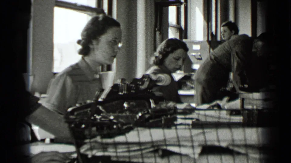 Several women are visible in a 1939 New York office, seated at desks with manual typewriters, papers, and tall windows behind them.