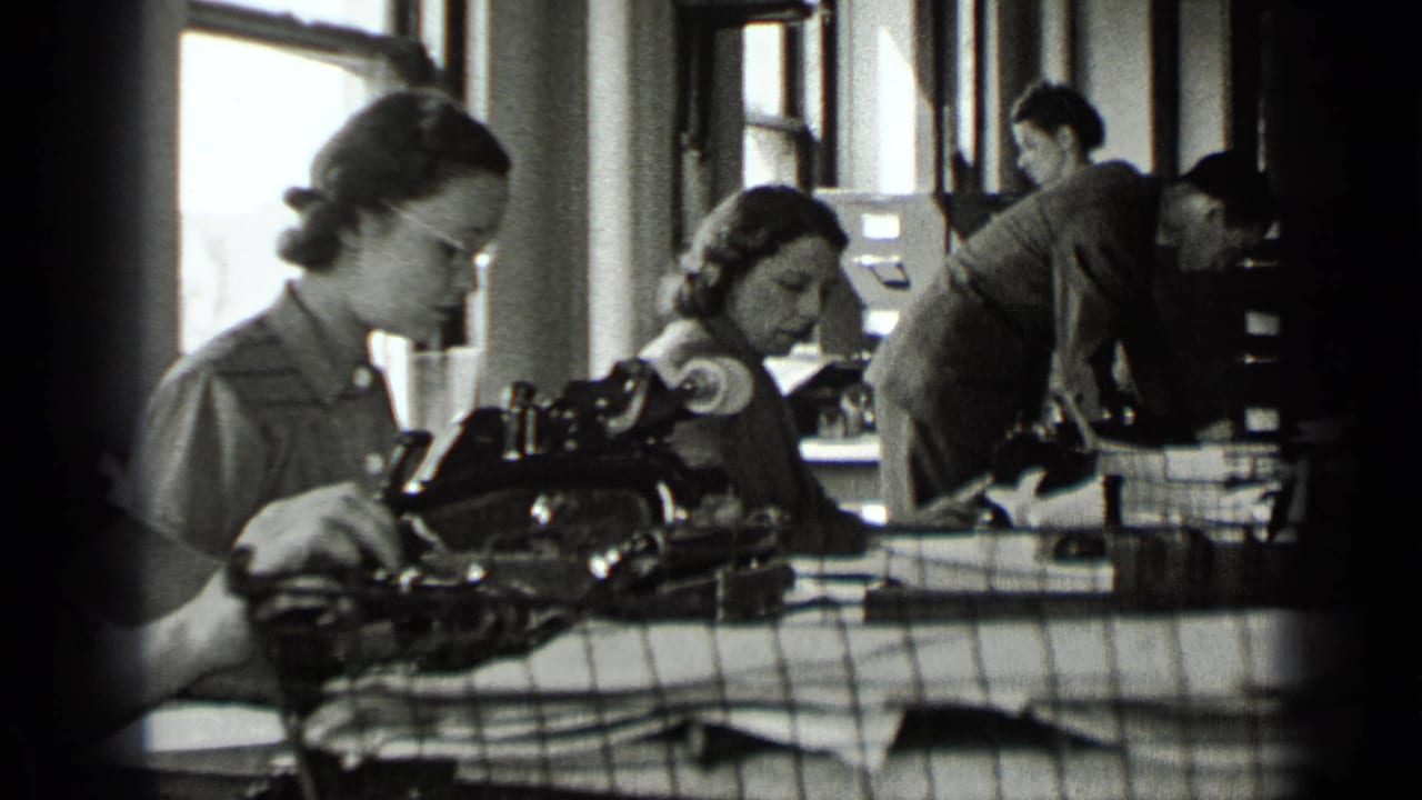 Women seated at typewriters in an office with a man working behind them.