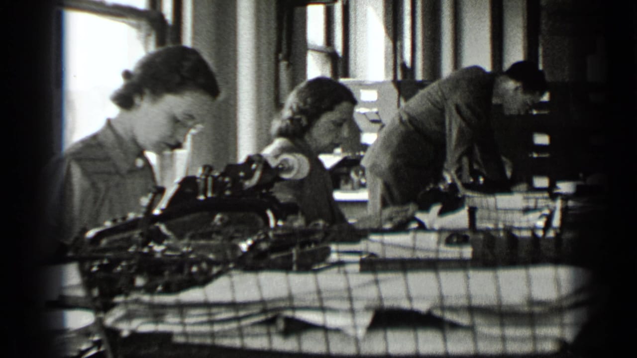 Two women work at typewriters while a man bends over a desk behind them.