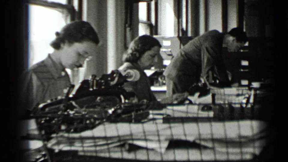 Women in a New York office type at desks as a suited man leans over work near cabinets and tall windows.