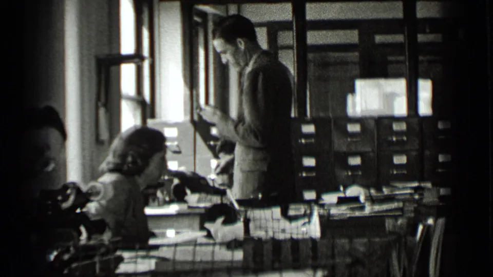 A man in a suit stands beside women working at typewriters in a New York office, with windows and cabinets behind them.