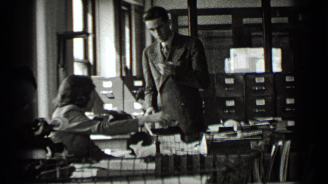 Office workers at desks with a standing man reviewing papers near filing cabinets.