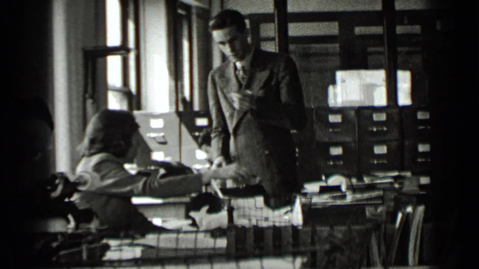 1939 New York office scene with a suited man standing by desks as women work with papers, typewriters, and filing cabinets.