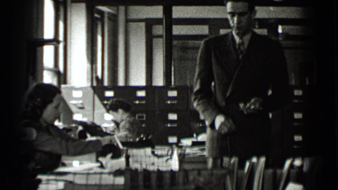 Man in suit faces the camera while clerks work at desks beside filing cabinets.