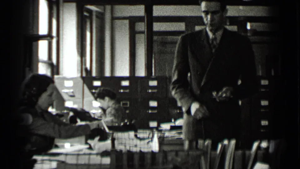1939 New York office interior with a suited man facing forward and women working at desks near card-index cabinets.