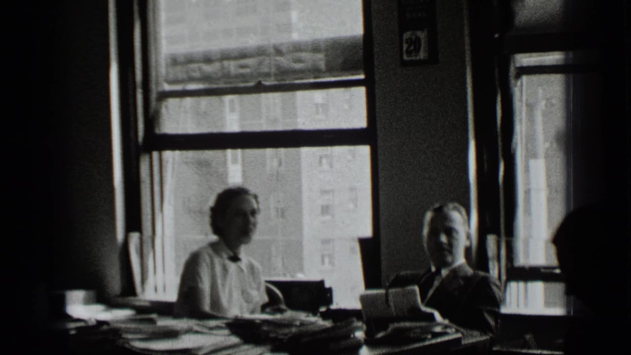 Man and woman seated at a cluttered office desk near large windows.
