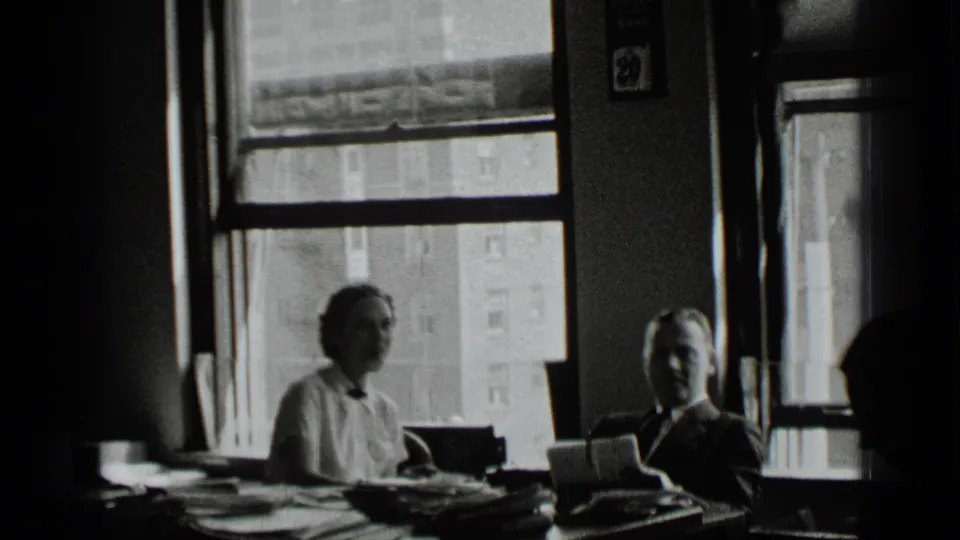 In a 1939 New York office scene, a man and woman sit behind a desk with paperwork and bright windows behind them.
