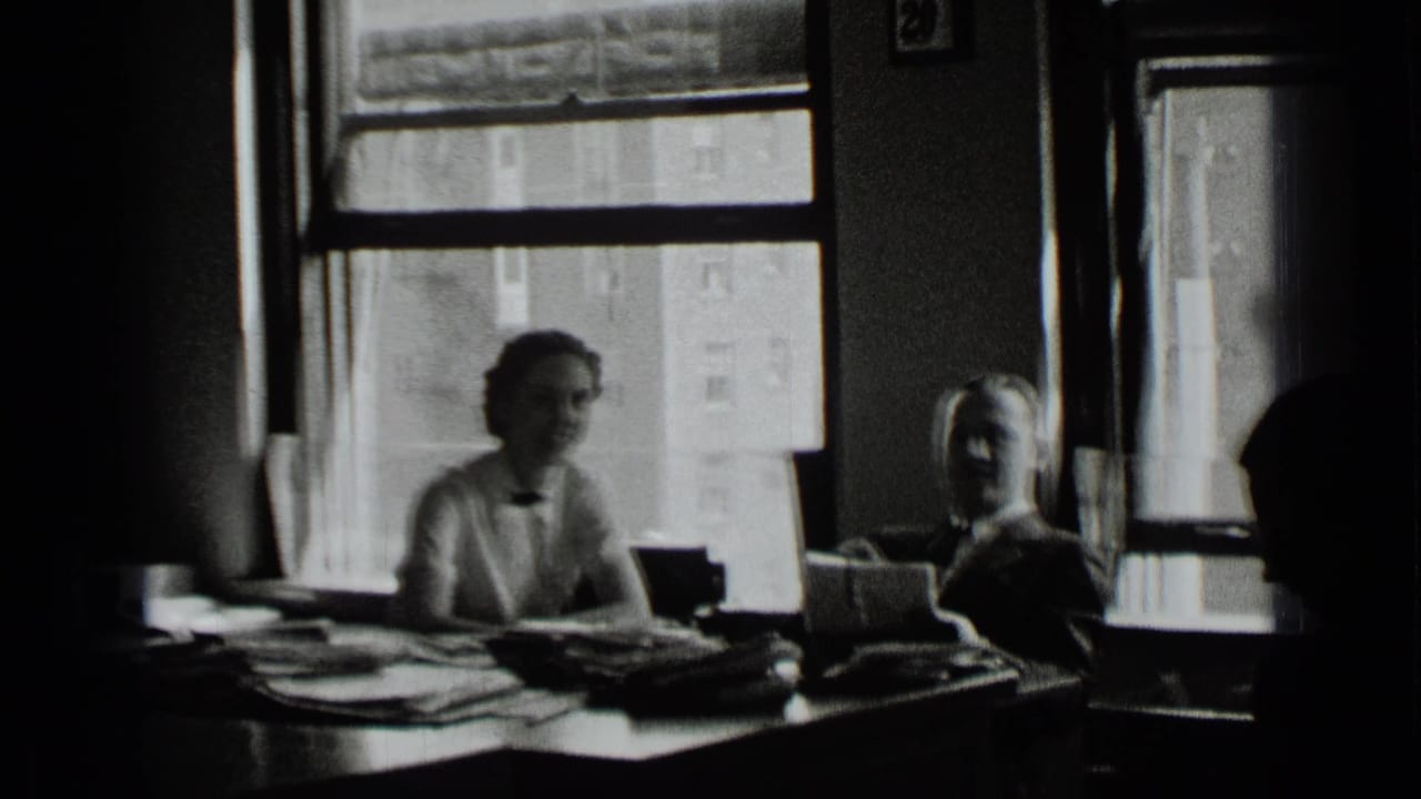 Two office workers sit behind a desk with papers and window light.
