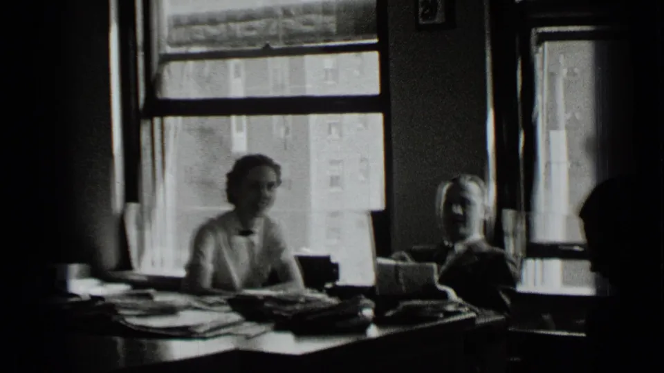 The man and woman remain seated at the office desk, with stacked documents and large New York windows visible behind them.
