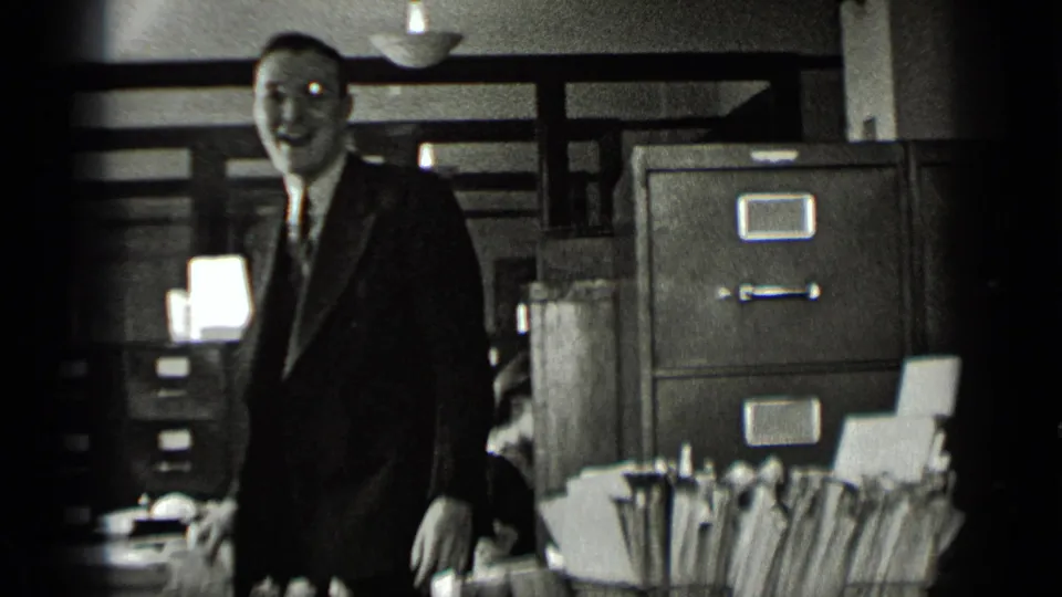 A suited man with glasses smiles near metal filing cabinets and a foreground row of folders in the office.