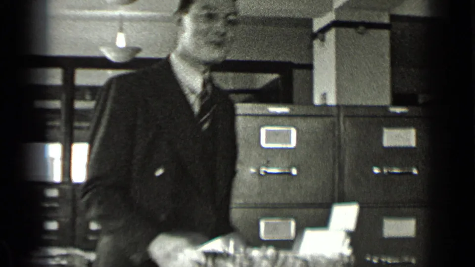 A suited male office worker with glasses stands close to metal filing cabinets, with folders and papers in the foreground.