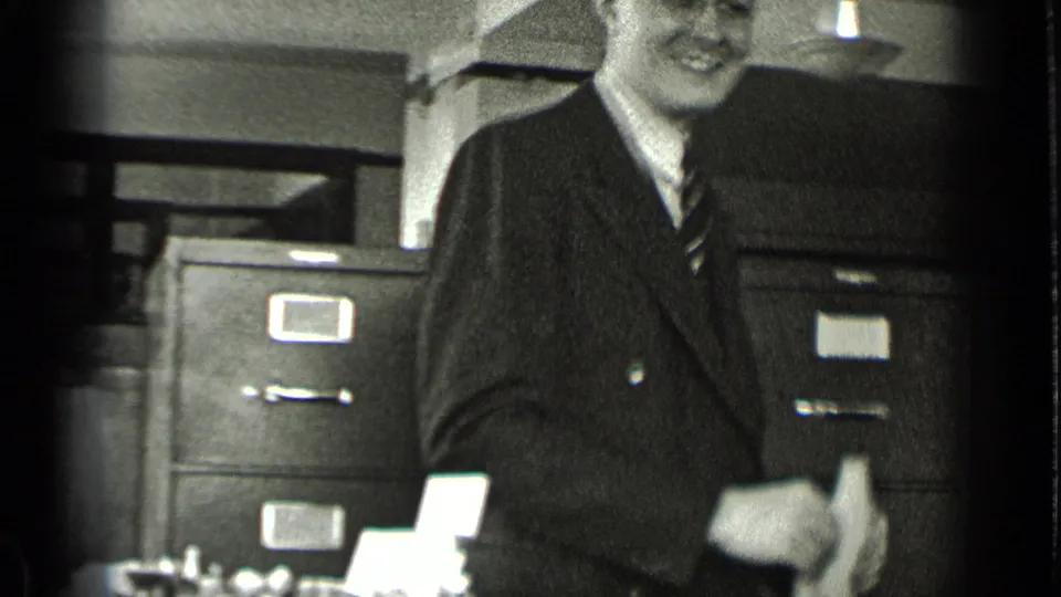 In a 1939 New York office scene, a suited man stands near filing cabinets with papers or desk items in the foreground.