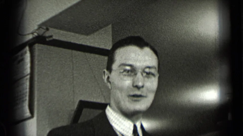A suited man with glasses appears to speak in a New York office interior, with paneled walls and furniture behind him.