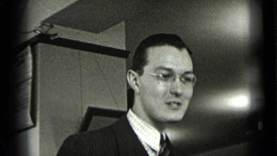 Close 8mm view of a suited man with glasses in the office, his face turned slightly downward as he appears to talk.