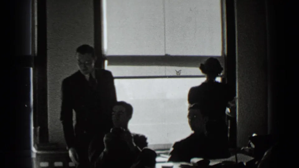1939 New York office interior with suited figures seated at desks and standing near large windows in high-contrast silhouette.