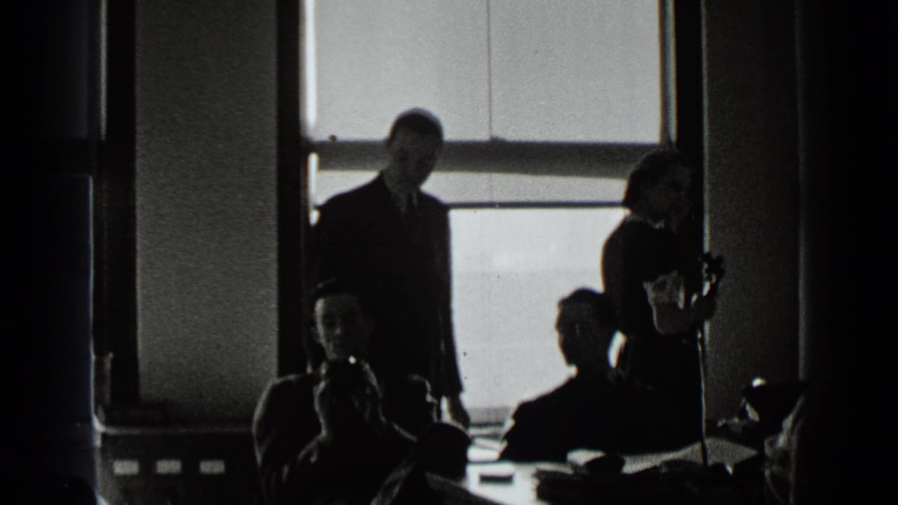 Men in suits and a woman near a telephone stand around desks by windows.
