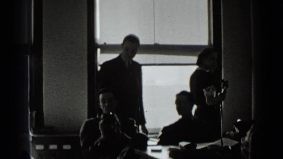 1939 New York office scene with suited men, seated figures, and a woman near a telephone beside bright windows.