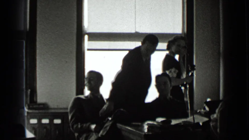 1939 New York office interior showing seated coworkers, a leaning suited man, and a woman holding a telephone near the window.