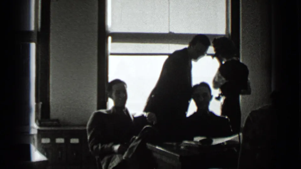 High-contrast 1939 New York office frame with silhouetted coworkers, desks, and a woman using or holding a telephone.