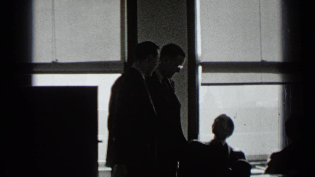 Two suited men stand beside seated figures in front of bright office windows.