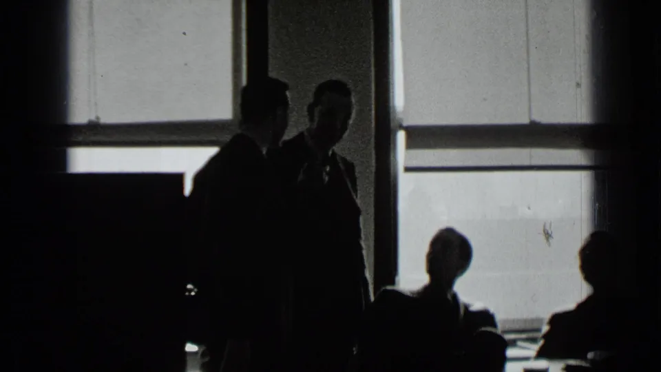 Silhouetted men in suits appear to speak in a dim New York office interior with seated adults nearby.