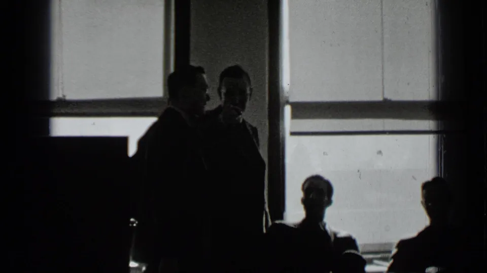 The frame shows a backlit office-like room with suited adults standing and sitting in shadow.