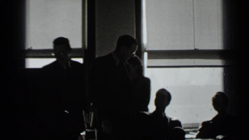 A dim 1939 New York meeting scene shows suited adults in silhouette near broad office windows.