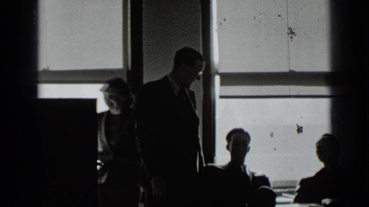 Silhouetted adults in an office near tall windows, with one suited man standing.