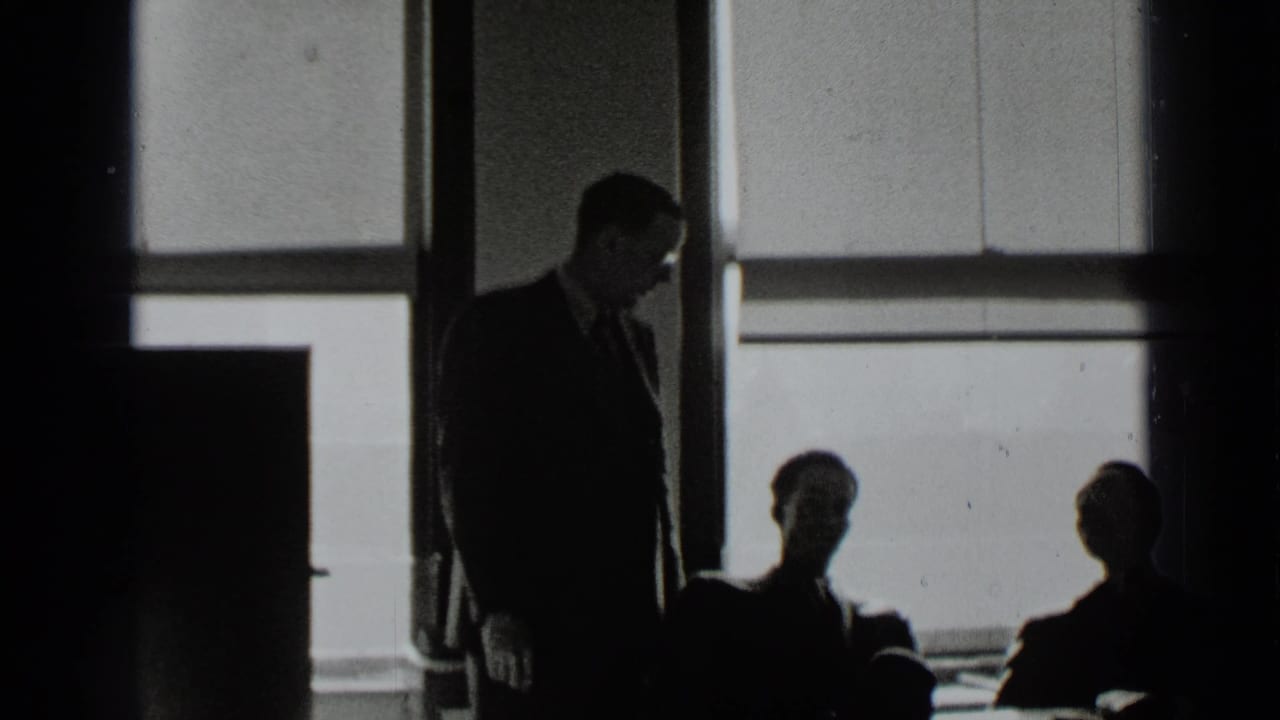 A suited man stands beside seated silhouettes in front of large office windows.