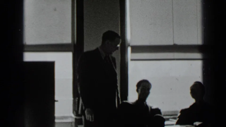 Backlit 1939 New York office scene with a suited man standing and seated adults silhouetted against tall windows.