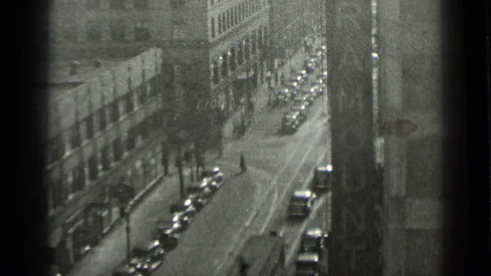 1939 New York home-movie frame looking down a city thoroughfare with vintage cars, sidewalks, and distant pedestrians.