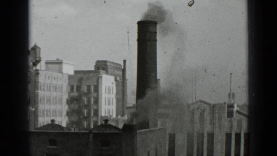 1939 New York 8mm frame showing a tall smokestack releasing dark smoke over brick and masonry city buildings.