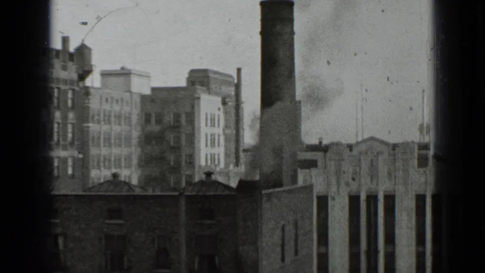 1939 New York 8mm frame showing an industrial city block with brick buildings, a smokestack, rooftop details, and smoke near the chimney base.