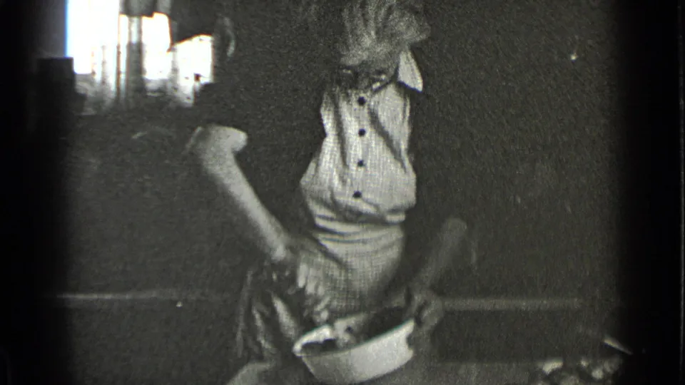 1939 8mm interior frame showing a woman in a buttoned blouse and apron mixing ingredients in a bowl in a dim kitchen or workroom.