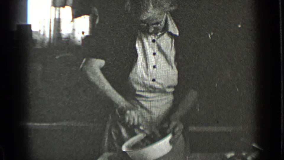 In 1939 New York home-movie footage, a woman in a light apron works over a white bowl in a dim interior.