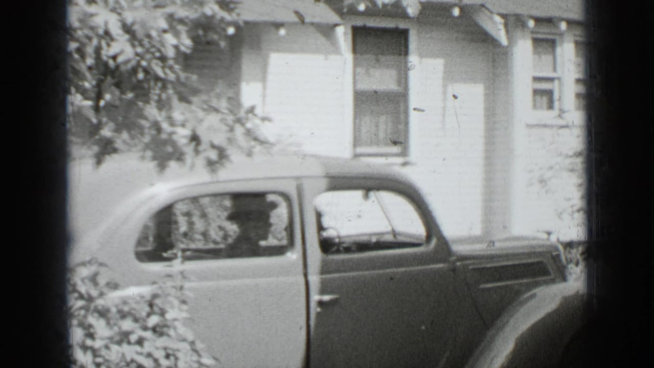Rounded 1930s sedan stopped outside a house with two adults visible inside.