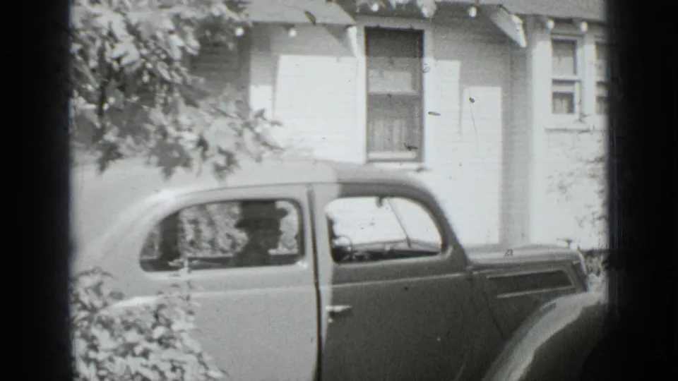 1939 New York home-movie frame of a rounded sedan beside a house, with a driver and passenger faintly visible through the windows.