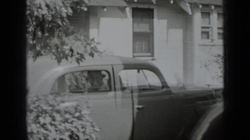 The sedan remains beside the New York residence, with the side windows, rear fender, house windows, and overhanging leaves visible.
