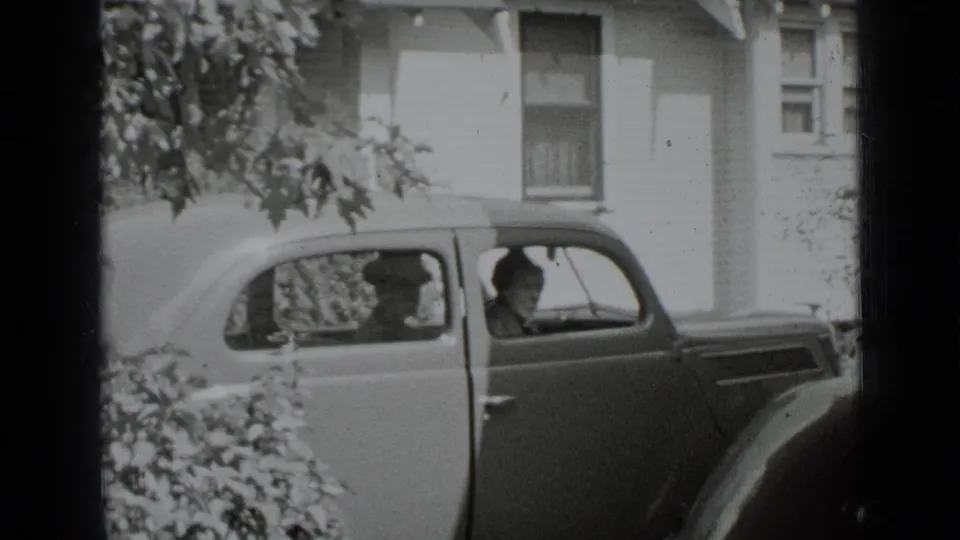 A man and woman appear inside the 1930s sedan, seen through the side windows in black-and-white home-movie footage.