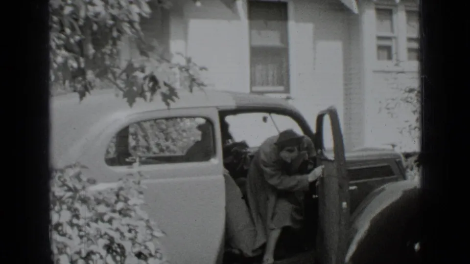 A woman in a coat and hat leans out of the sedan with the door open, while the driver remains seated outside the New York house.